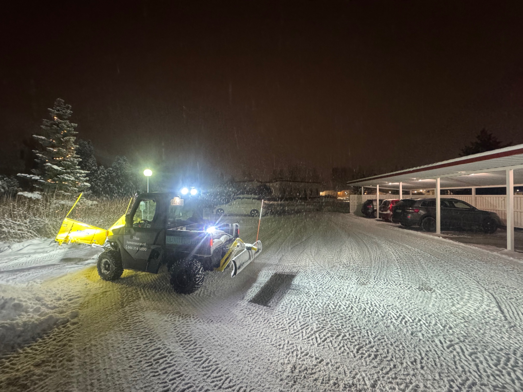 Luminurtsin aurauskone auraamassa autokatoksen edustaa yöllä lumisateessa